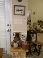 Small wood TV table against wall holding a large brown vase, vintage iron, decorative balls, and metal container. Two small cushioned wooden stools nearby. Light switch and plugged items on the wall.