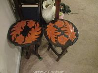 Pair of small wooden tables with round tops featuring carved orange floral patterns on a black background. Tables have dark wood foldable legs.