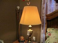 Brass table lamp with pleated cream shade placed on wooden nightstand near bed and window with lace curtains.