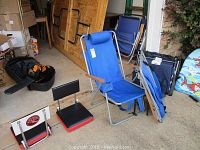 Three light blue lawn chairs with metal frames and padded headrests, two black and red cushioned stadium seats, dark blue folding stadium chairs stacked in the corner, and two colorful foam water floats with cartoon sharks leaning against the wall outdoors.