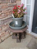 Small octagonal wood table with pot of pink and red flowering plants on top, sitting on concrete floor near stone wall.