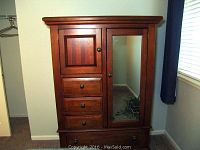 Front view of wood armoire showing the mirrored door on the right, wooden paneled door on left, and three drawers below the paneled door with round knobs.