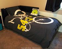 Twin bed frame with reversible corner headboard, black bedding with skeleton bike graphic, and yellow IKEA wall decor pieces on wall