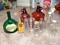 Ten assorted collectible glass bottles arranged on carpet, including amber Clorox bottles, green wine bottle, Minol container, and Crownford China bottles with ceramic stoppers.