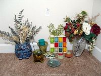 View of decorative vases with faux greenery and floral arrangements, candle holders, coasters in wooden holder, and decorative metal trivet on carpeted floor.