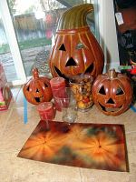 Three terracotta pumpkin candle lanterns, red candles, two glass vases (one filled with decorative dried botanicals), and part of metal wall hanging visible on floor.