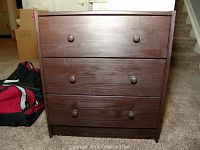 Front view of a small wooden dresser with three drawers and round wooden knobs. Surface shows some wear and scuffs.