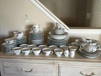 Full spread of the Nocturne China set arranged on a sideboard showing cups, bowls, plates, and serving pieces with floral design and silver trim.