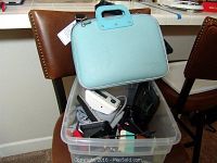 Clear plastic bin with assorted electronics and accessories including Cisco Linksys router, Omron blood pressure monitor, cordless phone base, phone cases, and chromatic pitch instrument. Blue SumacLife bag on top.