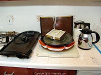 Photo of Rival electric griddle with thermostat, piled glass pizza stone, several trays, cutting boards, and two coffee pots on kitchen counter