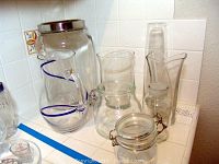 Glass pitchers, vases, and jar with lid arranged on a kitchen counter against white tiled backsplash.