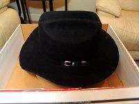 Black cowboy hat placed inside a tan-colored box, showing the front and side view with metallic buckle on the hatband.