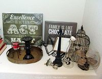 Side view of assorted home decor items on white shelf including wall signs, metal tray, candlestick holders, birdcage, glass door knobs, metal stands, and small metal fan.