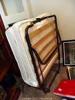 Folded twin size rollaway cot showing metal frame and mattress against a wall with some household items around.