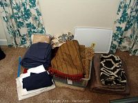 Assorted fabrics and linens arranged on floor including striped tassel fabric and animal print fabric next to plastic bin