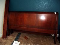 Wooden sleigh bed headboard and footboard in warm brown finish, leaning against a wall on carpeted floor.