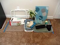 Wide view showing all lot items including Moen shower seat, canvas art, towels, baskets, bath mat, wire shelf, candle, and faux flowers.