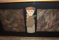 Framed shadow box display showing doll and two quilt samples with patchwork fabrics in earth tones hanging inside black frame