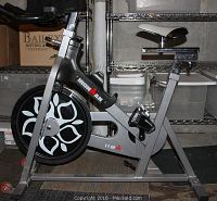 Side view of silver Ironman stationary bike with black and white flywheel, adjustable seat, and visible pedals.