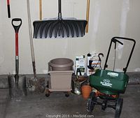 Photo showing the tools against the garage wall including metal shovels, black plastic shovel, green spreader, wheeled planter, brown pots, and garden chemical containers.