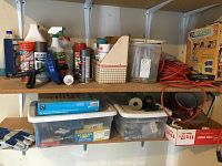 Photo showing two shelves with assorted spray paint cans, cleaning spray, small tools, orange electrical extension cord, and furniture moving pads.
