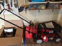 Red Troy-Bilt push lawn mower with attached black fabric grass catcher and two red gas cans beside it in a garage corner.