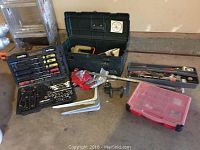 Overview of assorted tools spread on garage floor including plastic toolbox, socket set, plastic organizer boxes, hand tools, braces, and clamps