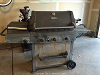 Full view of the Thermos gas grill showing its large metal body, control knobs, side shelves, wheels, and wear.