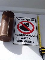 Photo showing one rusty cylindrical metal bucket next to a Neighborhood Watch Community metal sign with tape measure for size reference.