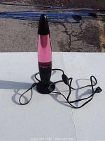 Pink lava lamp standing on white surface outdoors in sunlight with black base, pink liquid and wax inside, power cord visible.