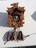 Front view of the wooden Swiss cuckoo clock showing carved leaves, bird on top, clock face with Roman numerals, and four pinecone weights with chains on a white background.