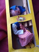 Photo of the Pelham Puppets Dutch Girl marionette puppet inside original yellow box, showing the puppet's head, bonnet, and upper body with strings.