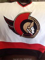 Front view of white Ottawa Senators hockey jersey showing team logo, red and black trim, and bottom hem detail.