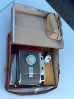 Open view of Polaroid camera inside brown leather carrying case with flash and pamphlets visible.