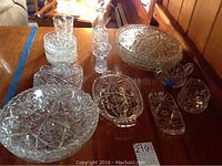 View of assortment of clear pressed and cut glass serving pieces on wood surface including serving plates, decanter with stopper, bowls, sugar and creamer.