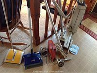 Overview photo of all items including vintage Electrolux canister vacuum, carpet sweepers, hand held vacuums, and accessories on floor next to wooden furniture.