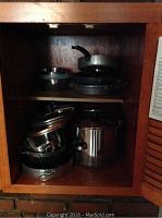 Kitchen cabinet with stacked pots, pans, and lids. Various sizes and styles including stainless steel and non-stick surfaces.