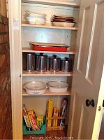 Cupboard shelf holding various kitchen items including two Corningware casserole pans, stainless steel canisters, a red enamel roaster, white casserole dishes, and kitchen wraps below