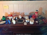 Long shot showing a variety of ceramic and glass decorative items arranged on a wooden sideboard. Items include vases, pitchers, figurines, and a lighthouse model.