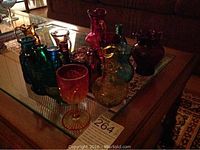 Photo showing a collection of colored glass items on a glass coffee table including various small vases, bottles, and a goblet in colors including red, green, blue, amber, and purple.