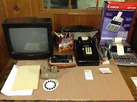 Photo shows full desktop assortment including small black Mitsubishi CRT TV, Canon MP11DX adding machine, black rotary phone, View-Master with reels, stapler, notepad, candy, and knife.