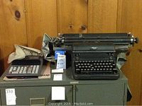 Vintage Royal typewriter and Casio adding machine positioned on a filing cabinet against a wood-paneled wall. A Kleenex box is also present beside the items.