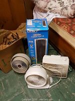 Four assorted fans and heater in box on green tiled floor near furniture.