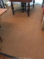 Image shows a large rectangular dark beige rug with a black border underneath wooden furniture, visible surface texture, and a slight discoloration or faded circle spot.