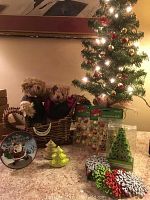 Two Russ teddy bears seated in a wicker basket sleigh, surrounded by Christmas decorations including a 24-inch illuminated tree, various ornaments in boxes, a decorative holiday plate, ceramic tree-shaped salt & pepper shakers, and snowflake coasters.