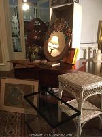 Photo showing hand carved floral mirror resting on wood furniture, wicker table with two shelves, and framed artwork on floor