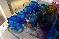 Top view showcasing eleven unique hand-blown glass cups in blue, two-toned, and mixed colors with decorative raised glass elements.
