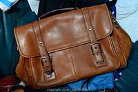 Brown leather satchel-style bag with two buckle straps and flap closure on a pile of cloth items.