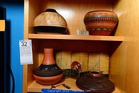 Wide shelf view showing four Navajo clay pots and two vintage bullets on upper shelf, lower shelf has a large shallow bowl and vase, with decorative screen in background.