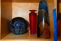 Three glass art pieces on a wooden shelf: a blue-green-amber elongated sculpture, a red translucent vase, and a black and blue glass sphere.